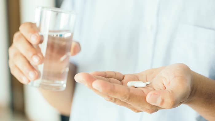 Een hand houdt een glas water vast en in de andere hand liggen witte pillen